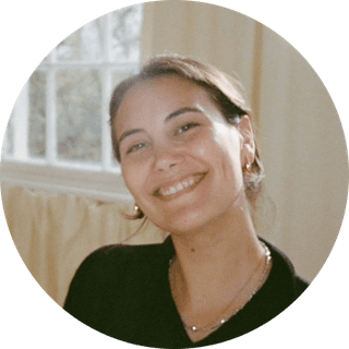 Smiling person with dark hair, wearing a black top and gold earrings, in a softly lit room with a window in the background.