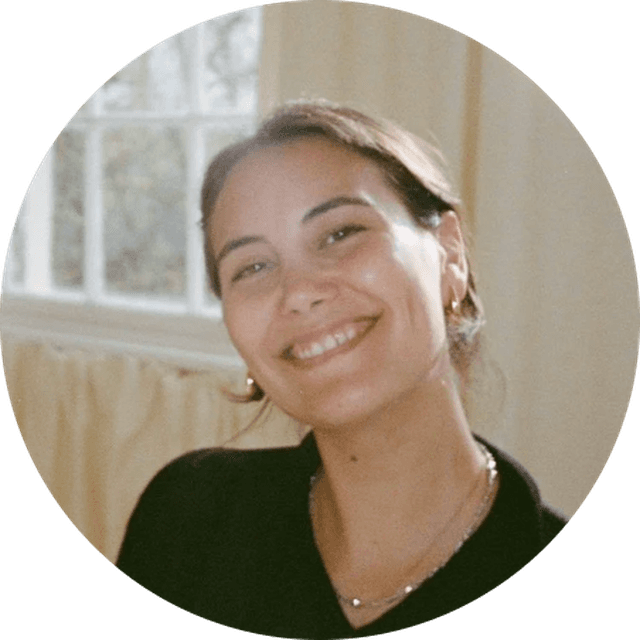 Smiling person with dark hair, wearing a black top and gold earrings, in a softly lit room with a window in the background.