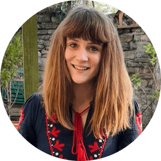 Smiling person with long brown hair, wearing a navy top with red embroidery, standing outside near a stone wall and greenery.