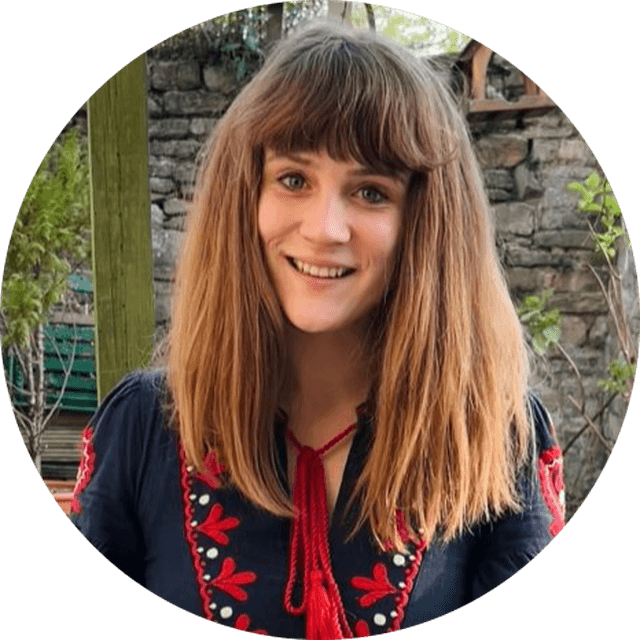 Smiling person with long brown hair, wearing a navy top with red embroidery, standing outside near a stone wall and greenery.
