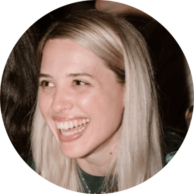 A person with long blonde hair smiling brightly, wearing a dark top, against a blurred background.