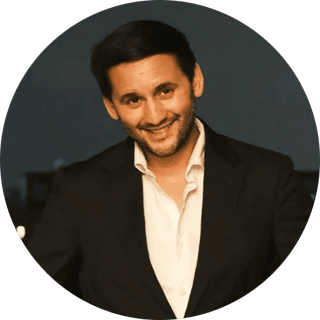 Smiling dark-haired man in a black suit and open-collar white shirt, head-and-shoulders portrait on a dark background.