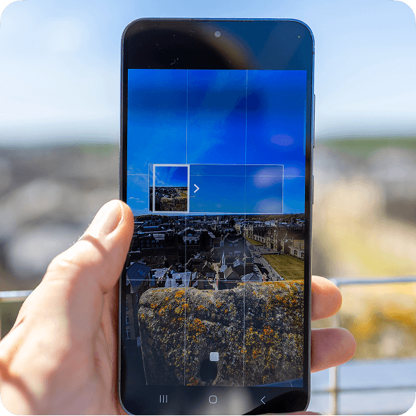 A hand holds a smartphone displaying panorama mode, with a cityscape image on the screen. The background is blurred.