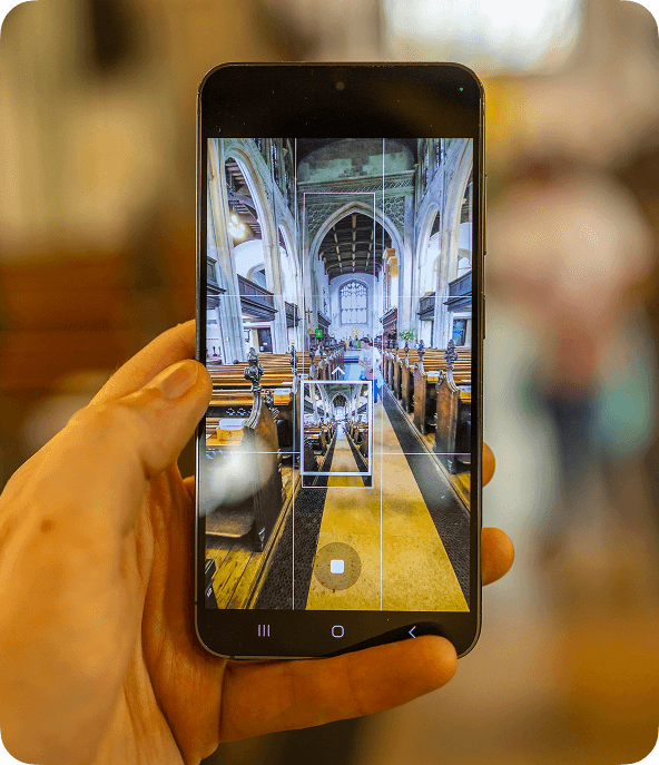 Hand holding a smartphone capturing an interior view of a church with arches, pews, and stained glass in the background.