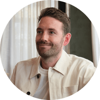 Smiling man with short hair and beard, wearing a light jacket and white shirt, seated indoors with sheer curtains in the background.