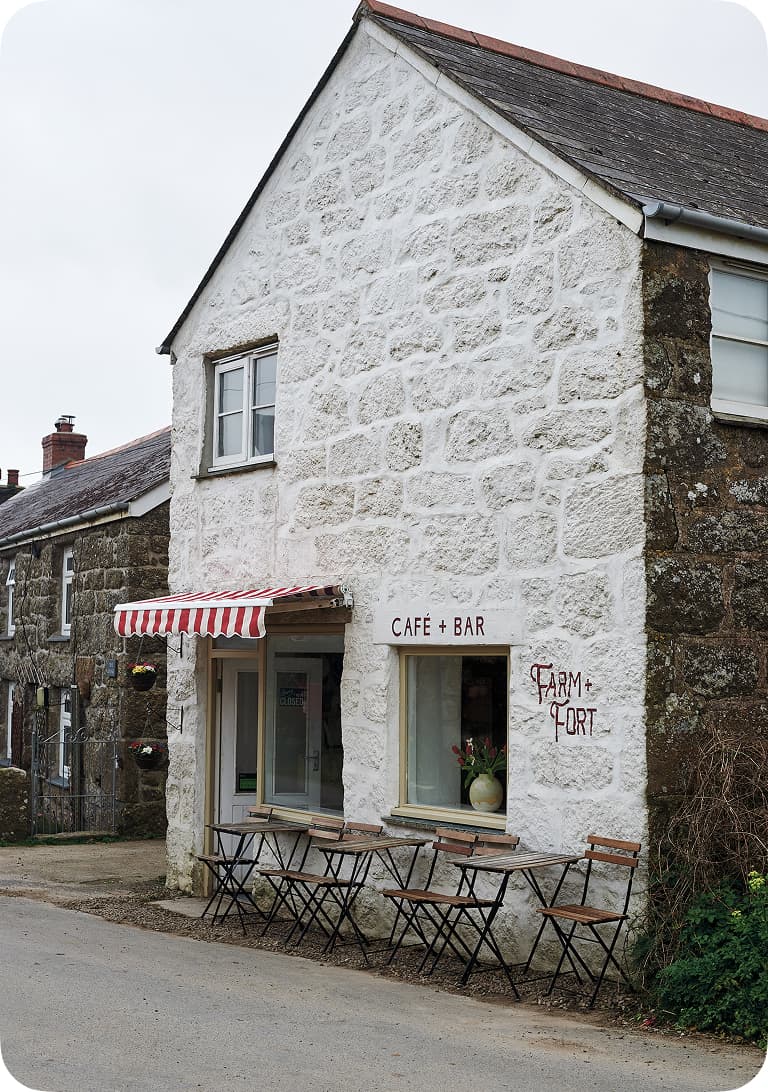 Stone building with a "Café + Bar" sign, red-striped awning, and outdoor seating. A "Farm Fort" sign is painted on the wall.