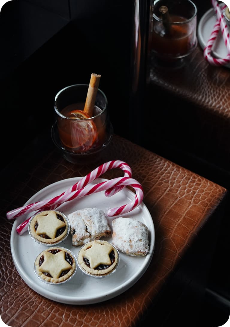 Plate with star-topped mince pies, powdered sugar pastries, and candy canes. Glass with spiced drink and cinnamon stick nearby.