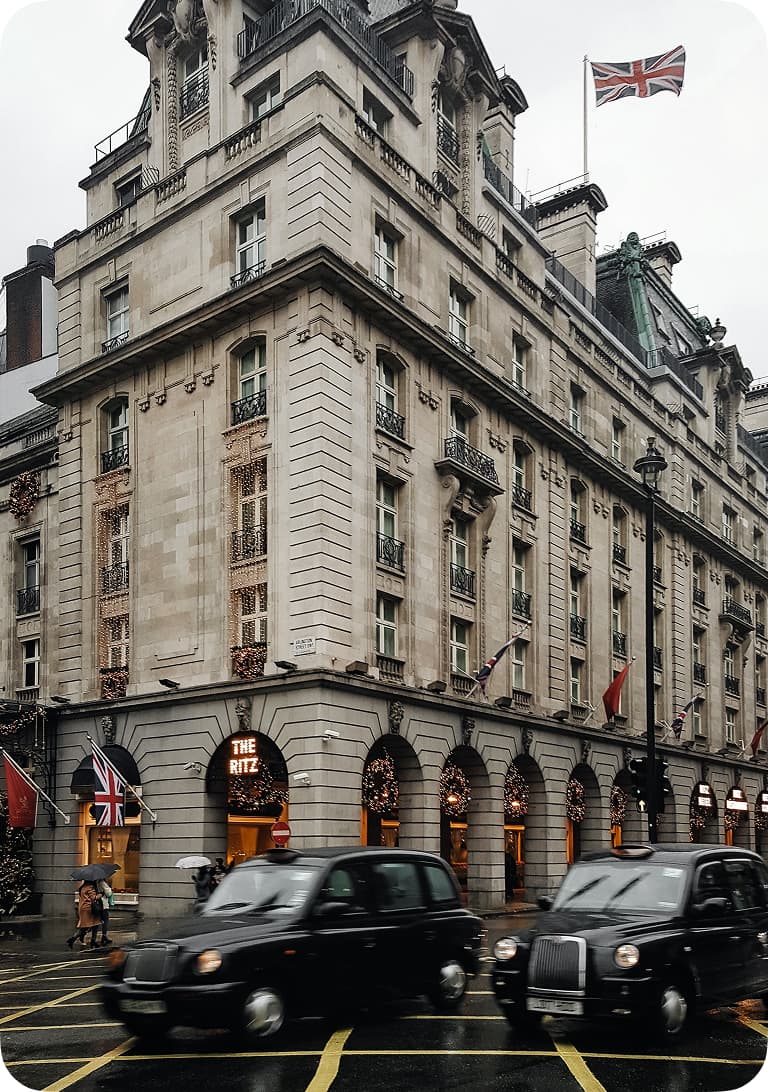 The Ritz, a historic building with arched windows, a British flag, and taxis passing by on a rainy day.