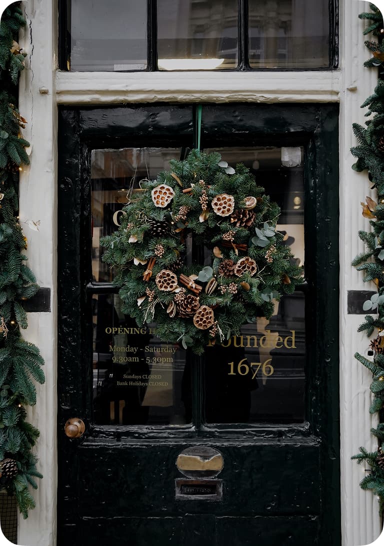 A festive wreath with pinecones and dried elements hangs on a dark green door, flanked by garlands, with gold text detailing store hours.