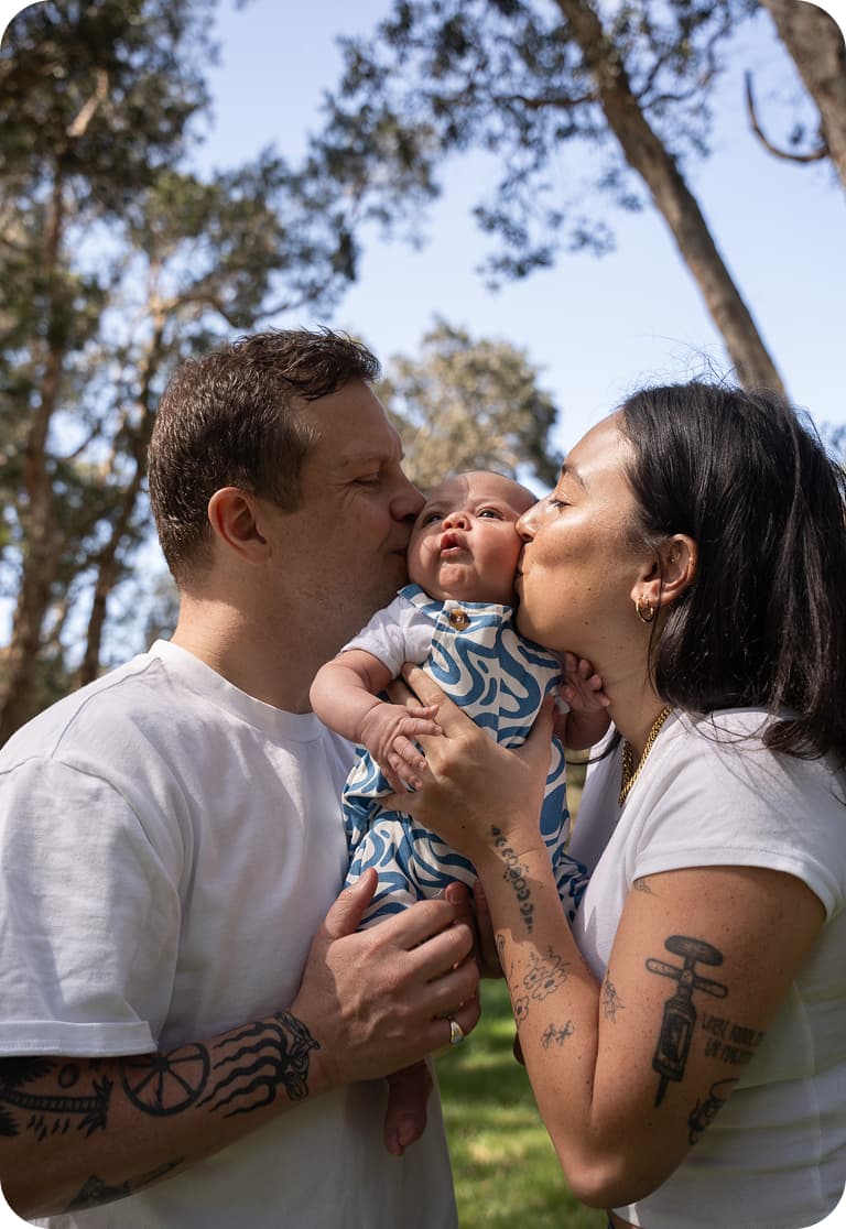A couple lovingly kisses their baby, who is wearing a blue patterned outfit, while standing outdoors surrounded by trees.