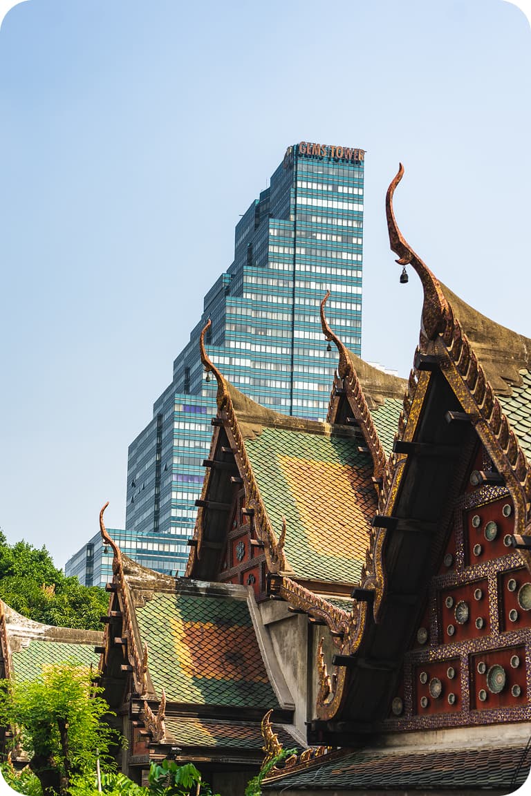 Ornate Thai temple roofs with gilded finials in foreground, modern stepped glass skyscraper rising behind under a clear sky.
