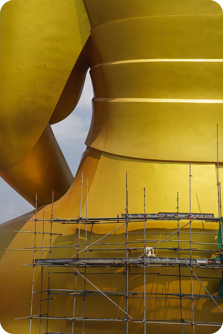 Close-up of a large golden statue's curved torso with metal scaffolding for maintenance against a pale sky.