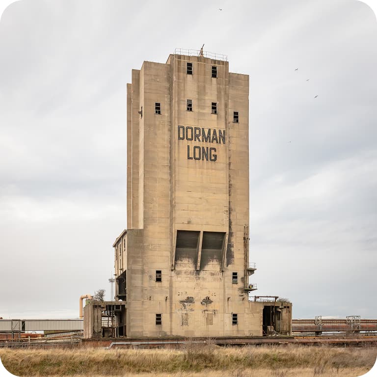 Tall, rectangular concrete building labeled "Dorman Long" against a cloudy sky, surrounded by industrial structures and grassy field.