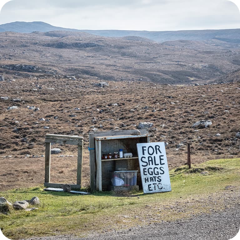 A rural roadside stand with a sign reading "For Sale Eggs Hats Etc" against a barren, rocky landscape under a cloudy sky.