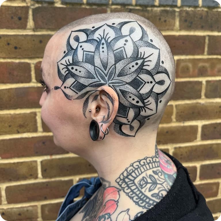 Person with a shaved head featuring an intricate mandala tattoo, wearing ear gauges and a black top, standing against a brick wall.