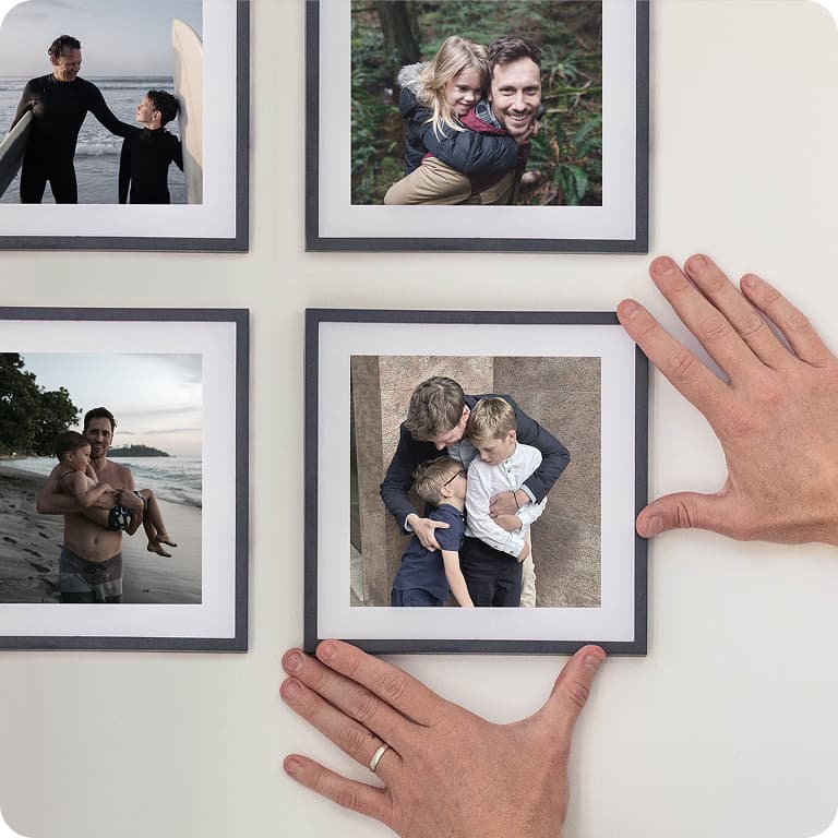 Hands adjusting a framed photo of a man and two boys, surrounded by other family pictures on a white wall.