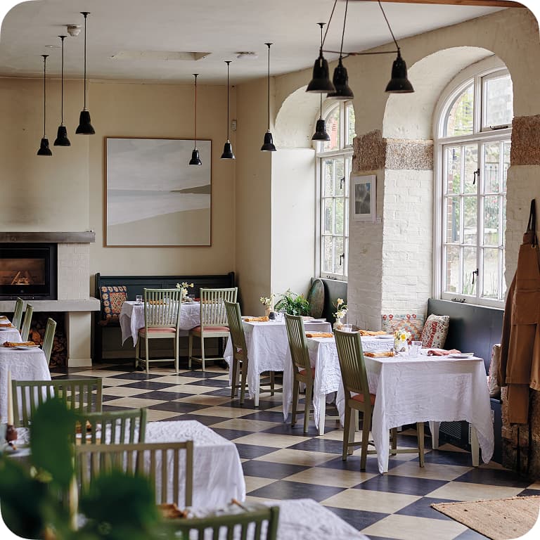 Cozy restaurant interior with checkered floor, white tablecloths, and large windows. Tables are set with dishes and flowers, and pendant lights hang above.