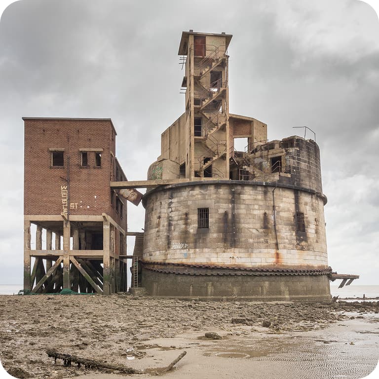 A weathered, round stone fort with an attached brick building on a muddy shore under a cloudy sky.