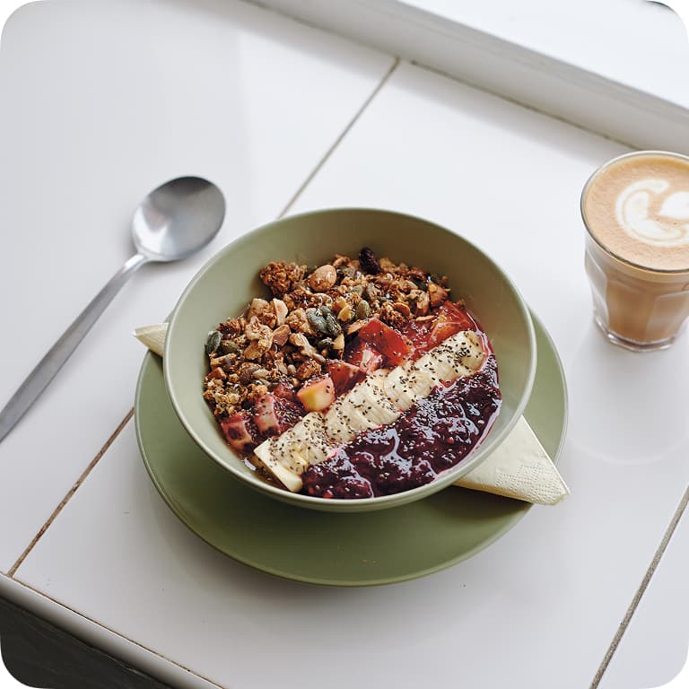 A bowl of granola with banana slices, berries, and seeds, next to a spoon and a cup of latte on a white table.