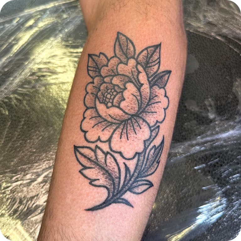 Black ink tattoo of a detailed flower with leaves on a person's forearm, placed on a reflective surface.