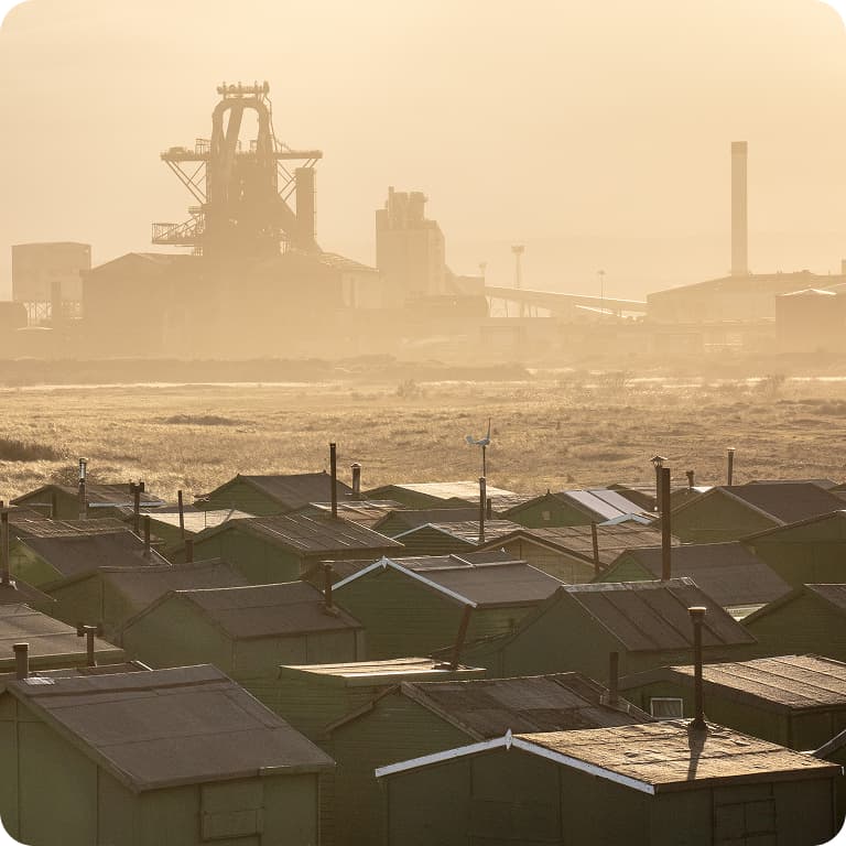 A hazy industrial landscape with a large factory in the background and rows of small green buildings in the foreground at sunset.