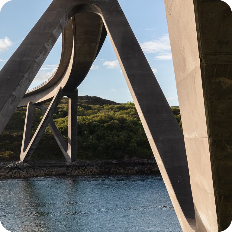 Curved concrete bridge over calm water with lush green hills in the background under a clear blue sky.