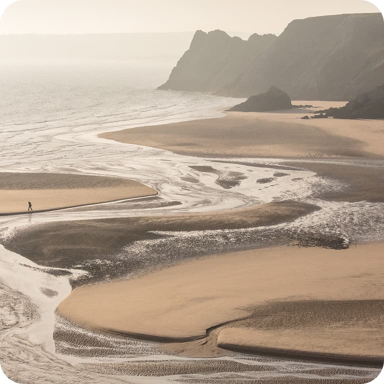 A lone person walks along a vast, winding beach with gentle waves and distant cliffs under a hazy sky.