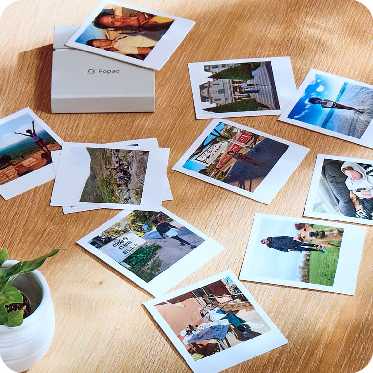 A collection of scattered Polaroid photos on a wooden table, with a small box nearby and a potted plant in the corner.