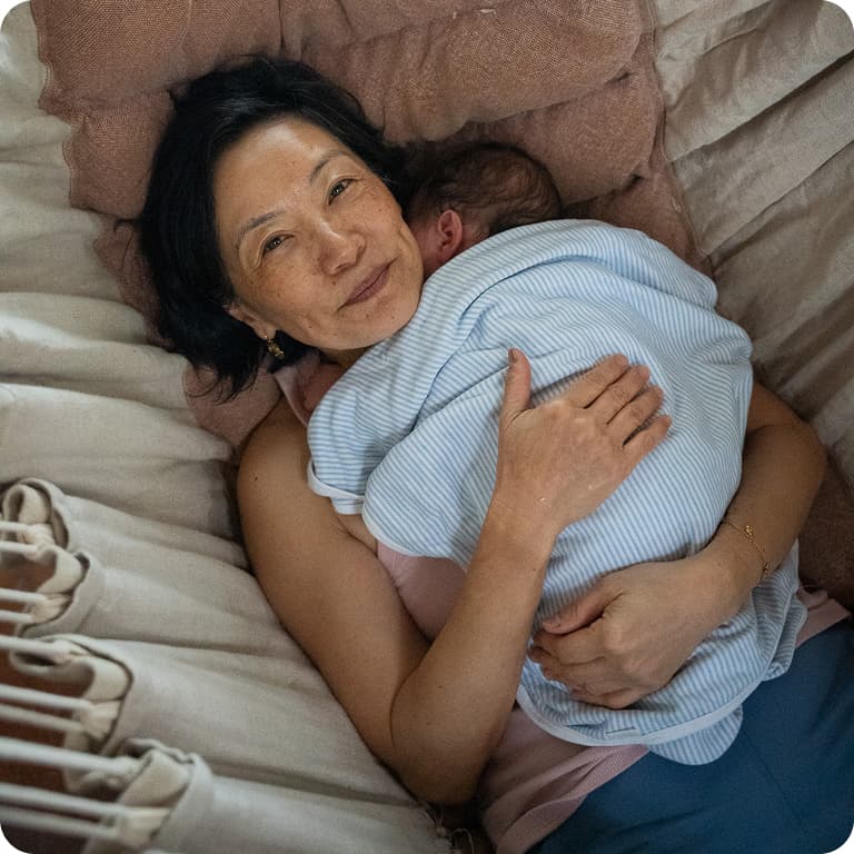 A woman lying on a bed, smiling while holding a baby wrapped in a blue blanket, surrounded by soft pillows.