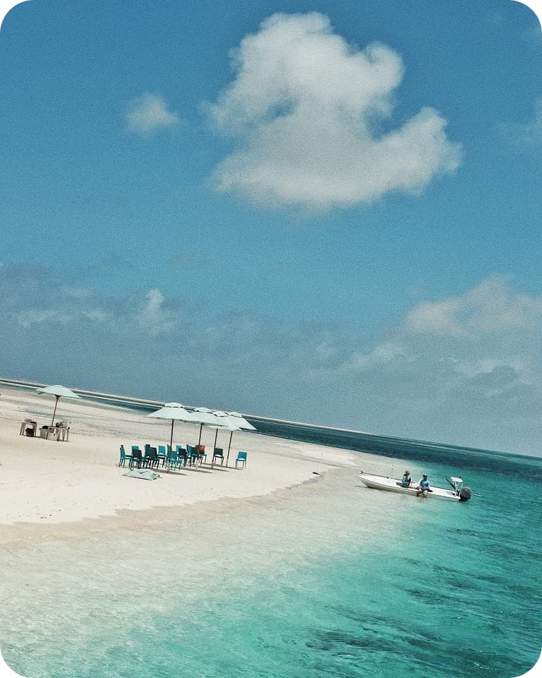 Tiny sandy islet with umbrellas and blue chairs, turquoise shallow water and a small motorboat with two people under a bright blue sky and lone cloud.