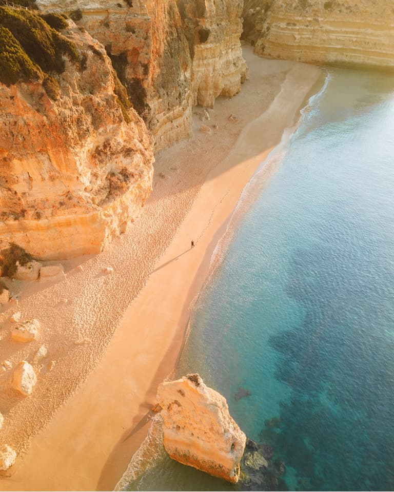 Aerial view of a serene beach with golden cliffs, clear turquoise water, and soft sand, bathed in warm sunlight.