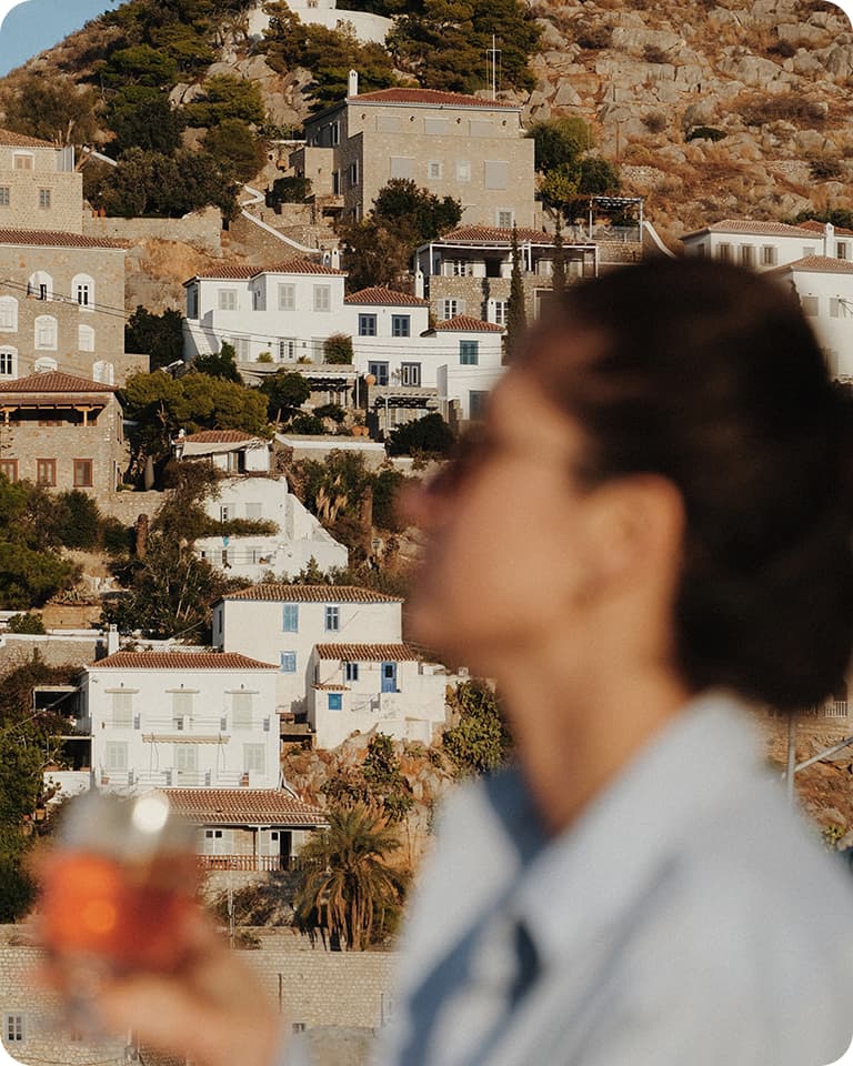 Blurred person holding a drink in foreground with sunlit hillside of white Mediterranean houses behind.