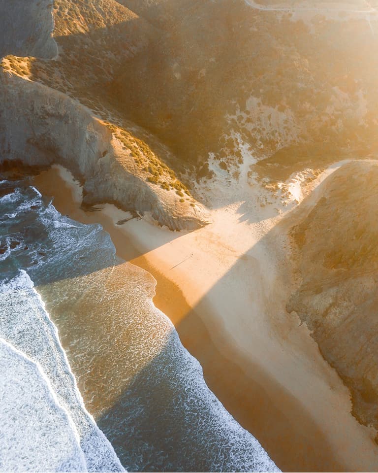 Aerial view of a secluded beach surrounded by rocky cliffs, with waves gently crashing on the shore and soft sunlight illuminating the scene.