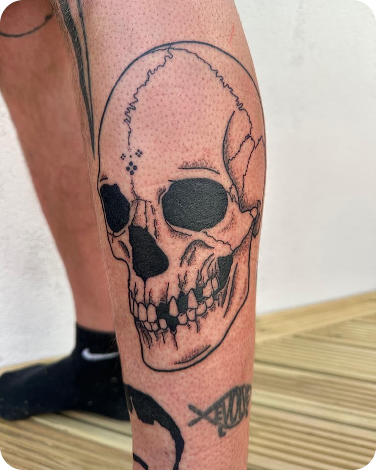 A detailed skull tattoo on a person's leg, featuring bold lines and shading. The leg is resting on a wooden surface.