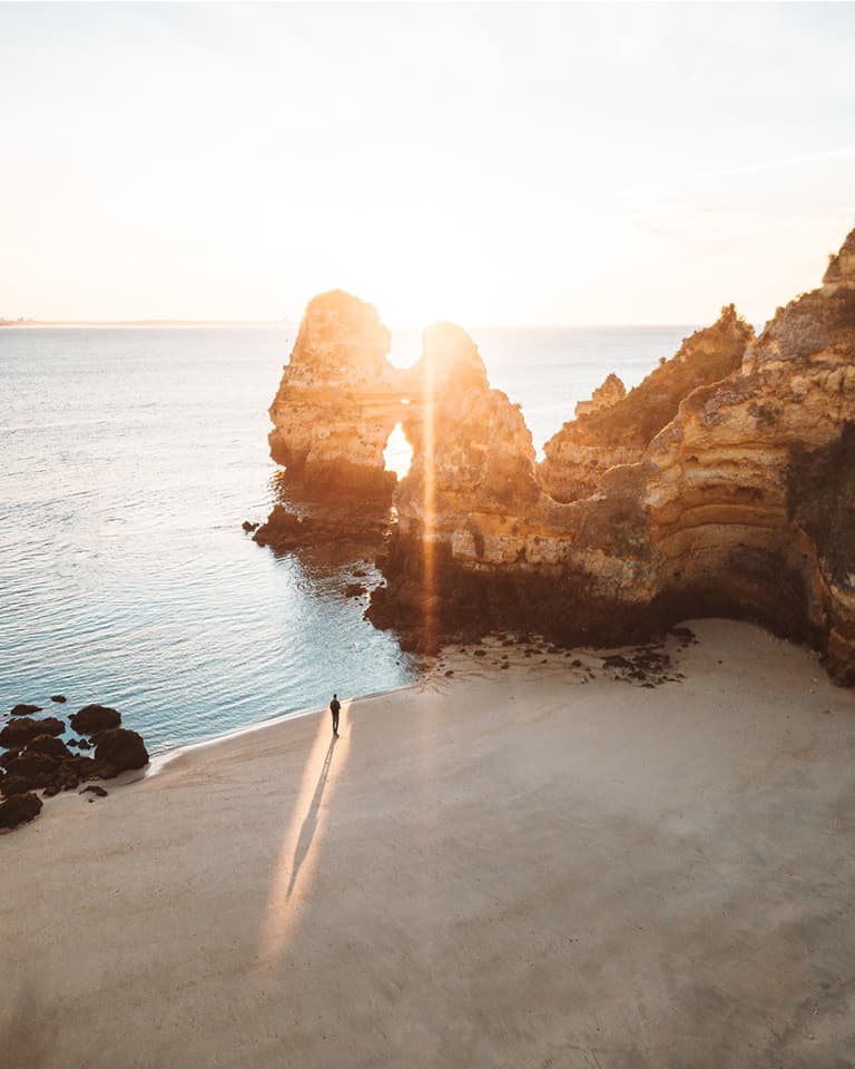 Person standing on a sandy beach at sunrise, with rock formations and the ocean in the background. Sunlight creates a long shadow.