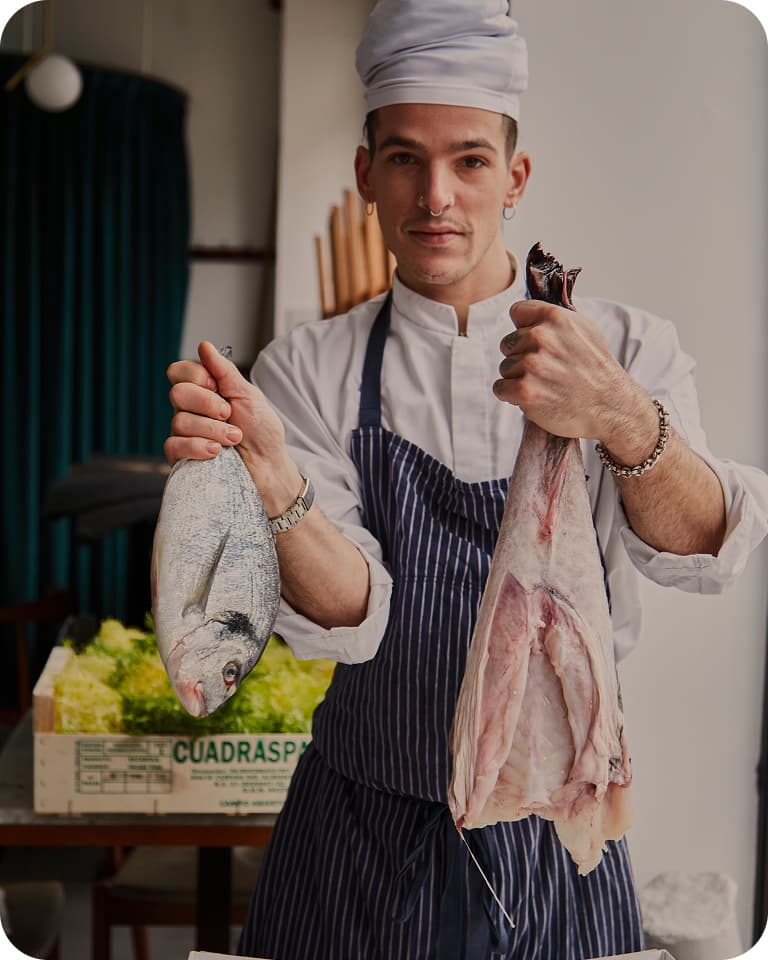 Chef in white hat and striped apron holds a whole fish in one hand and a gutted fish in the other, produce box behind him.