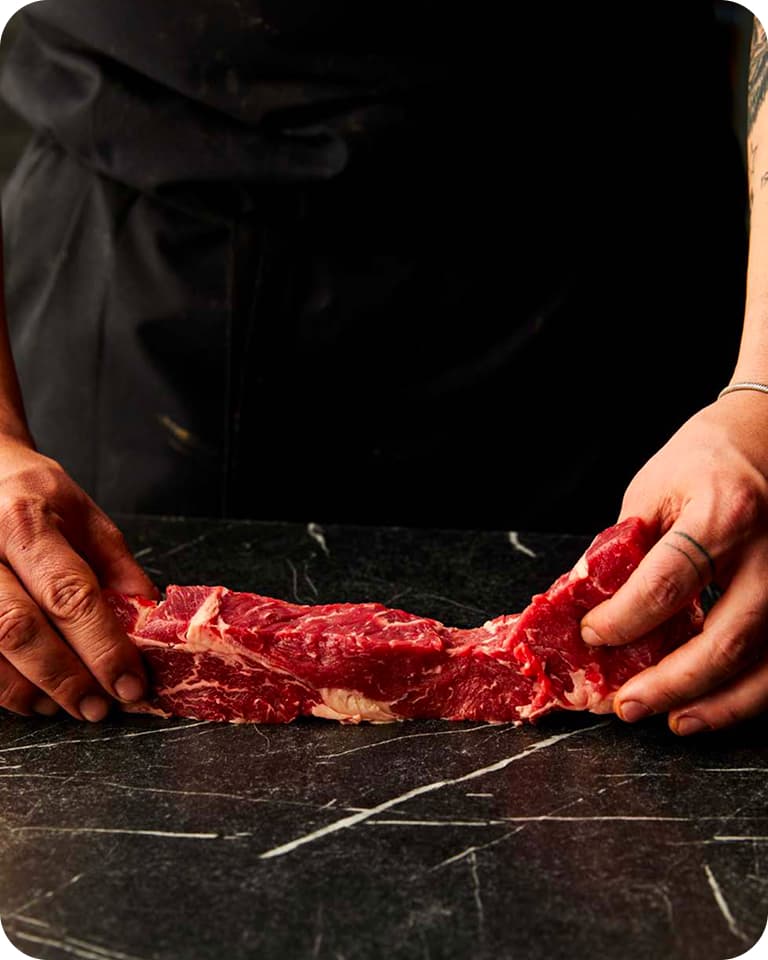 Cook's hands holding a long marbled strip of raw beef on a dark marble countertop.