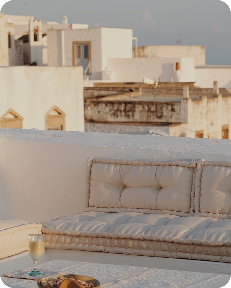 Sunlit rooftop terrace with padded beige cushions, a low table holding a glass of white wine and a small bowl, white Mediterranean buildings beyond.