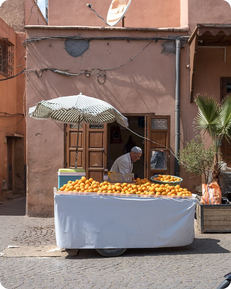 A street vendor sells oranges from a cart with a striped umbrella in front of a rustic building on a cobblestone street.