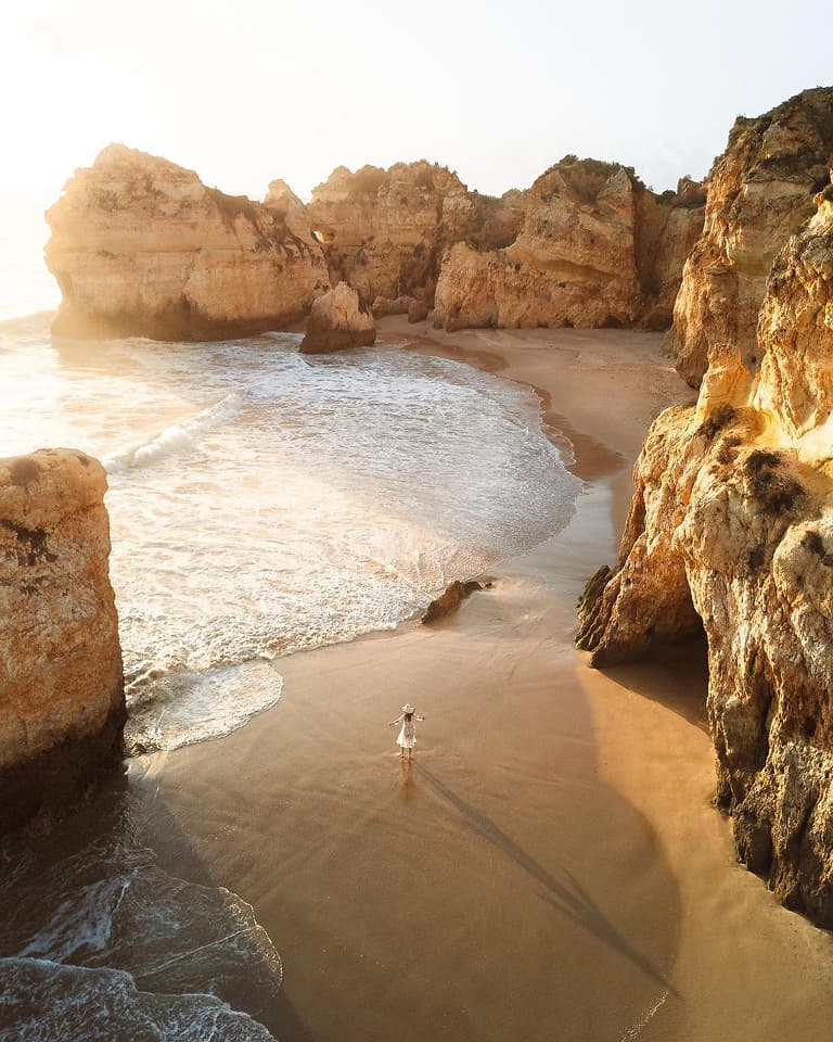 A person in white stands on a sandy beach surrounded by towering cliffs, with gentle waves lapping at the shore under a soft, glowing sky.