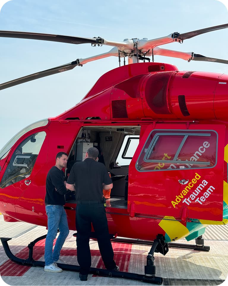 Two people stand by an open red helicopter marked "Advanced Trauma Team" on a helipad under a clear sky.