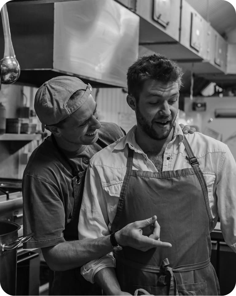 Black-and-white photo of two cooks in a kitchen; one playfully points at the other's apron while both smile.