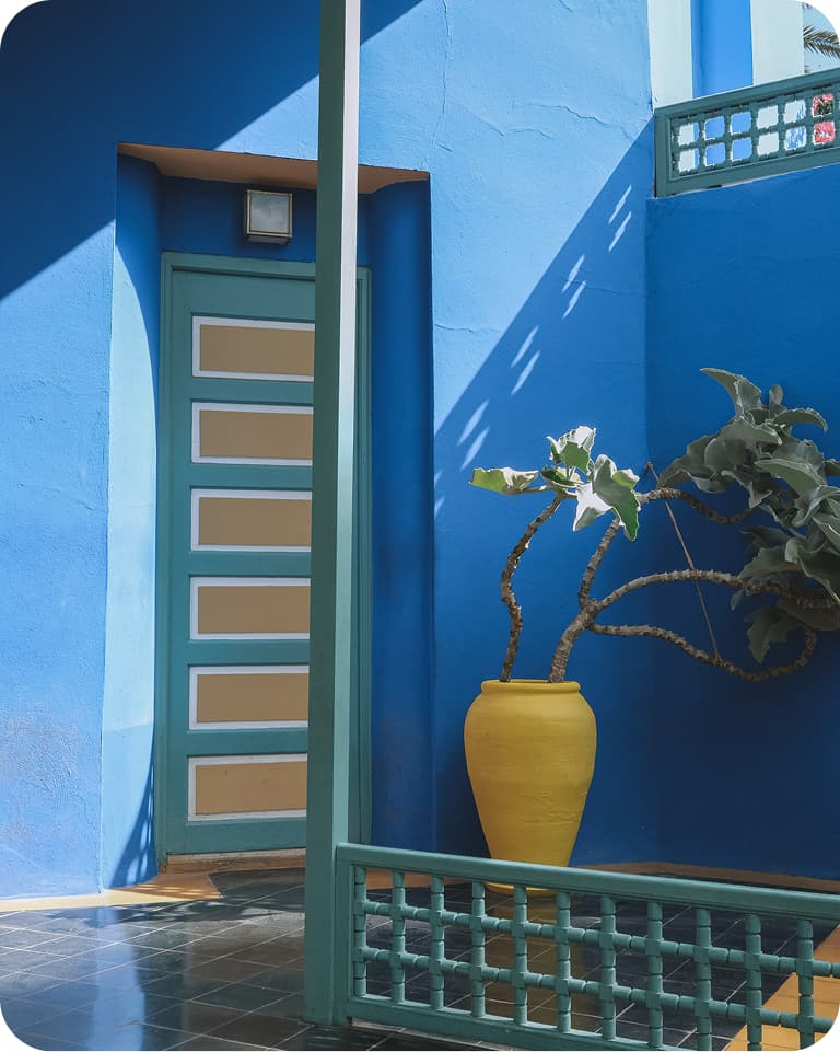 Bright blue courtyard with a striped door, yellow vase, and leafy plant. Sunlight casts shadows on the tiled floor and walls.