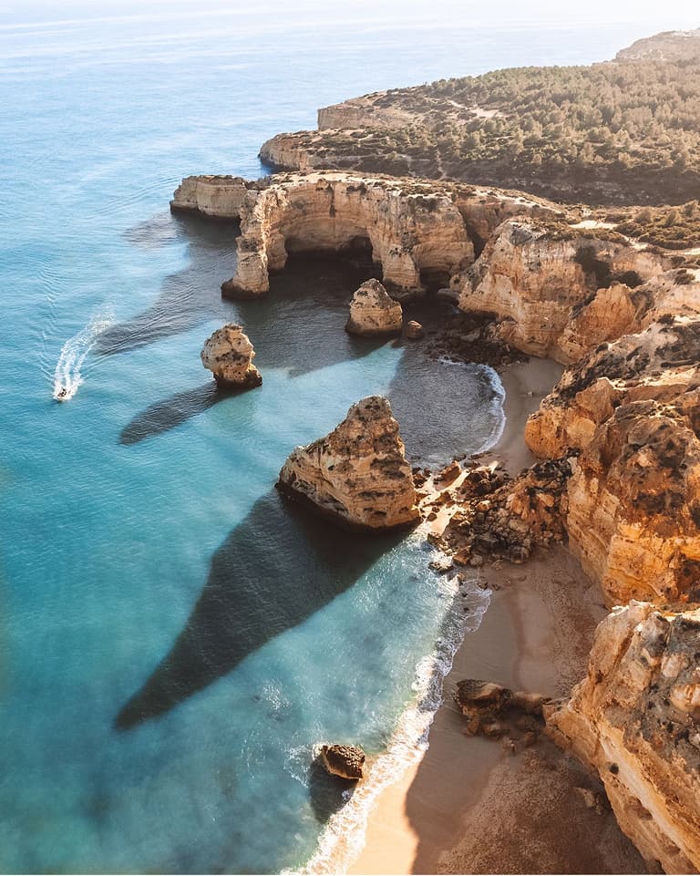 Drone photograph above rocky cliffs descending onto a golden sandy beach, with a boat speeding through the surrounding turquoise water