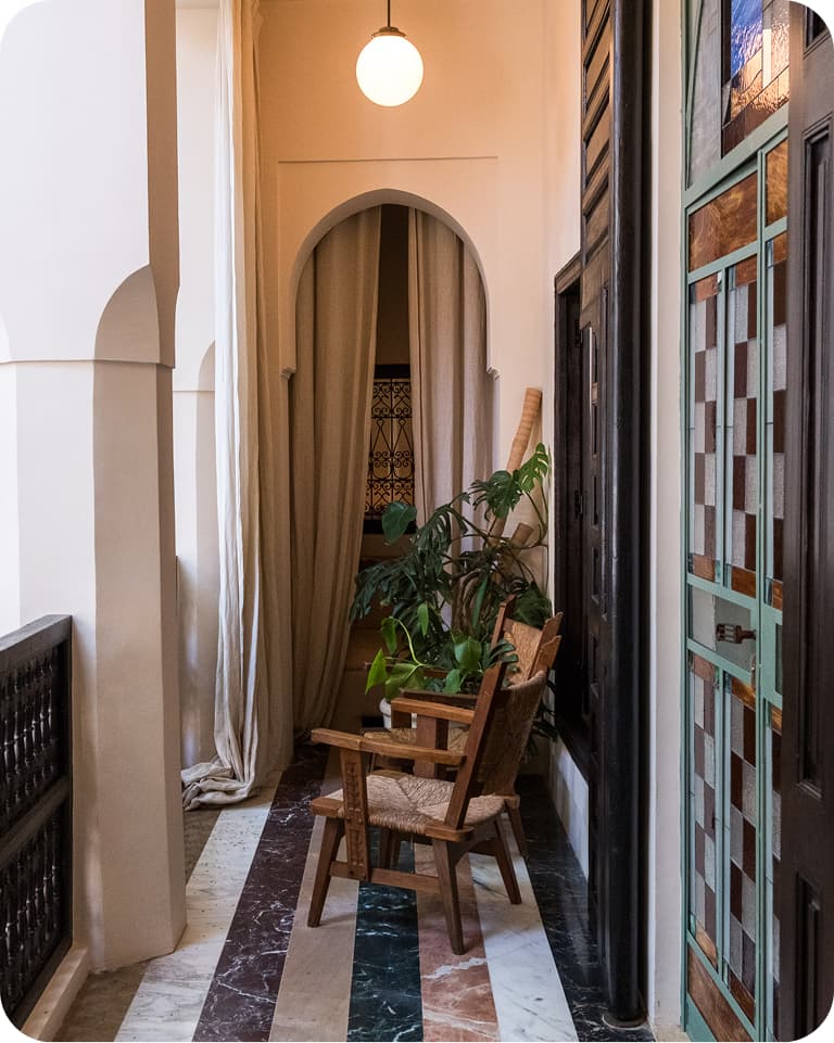 Cozy balcony with a wooden chair, potted plants, arched doorway, and colorful tiled floor, illuminated by a hanging light.
