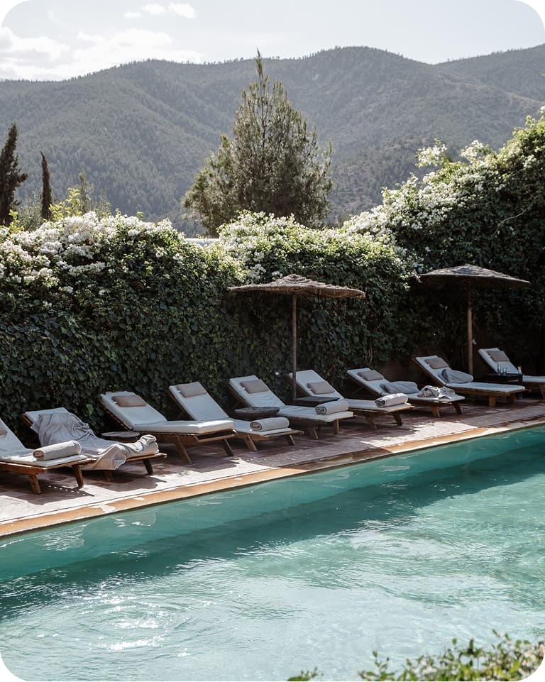 Poolside scene with lounge chairs and umbrellas, surrounded by lush greenery and mountain views under a clear sky.