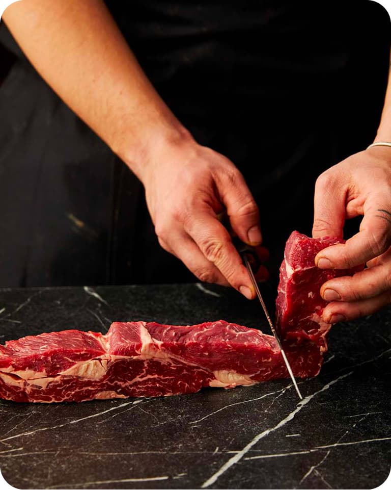 Hands slicing a raw strip of marbled beef with a knife on a dark marble countertop.