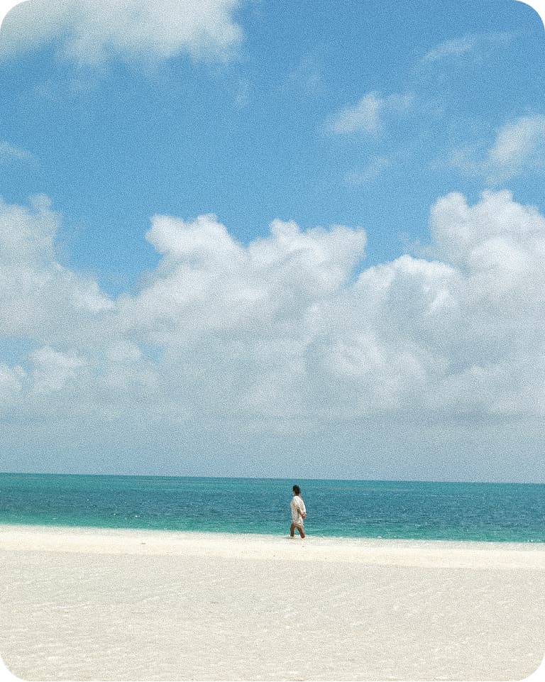 Solitary person walking along a white sandy beach by turquoise sea under a vast blue sky filled with fluffy clouds.