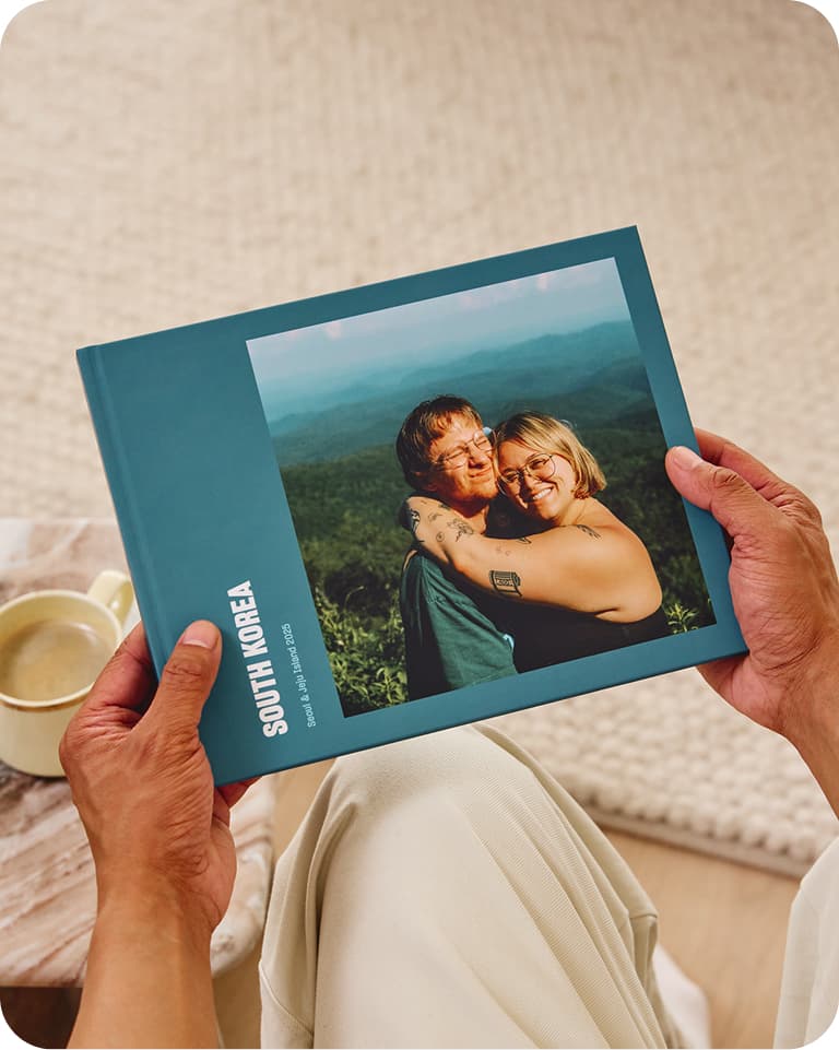 Person holding a photo book titled "South Korea" with a cover image of a smiling couple embracing against a scenic landscape.