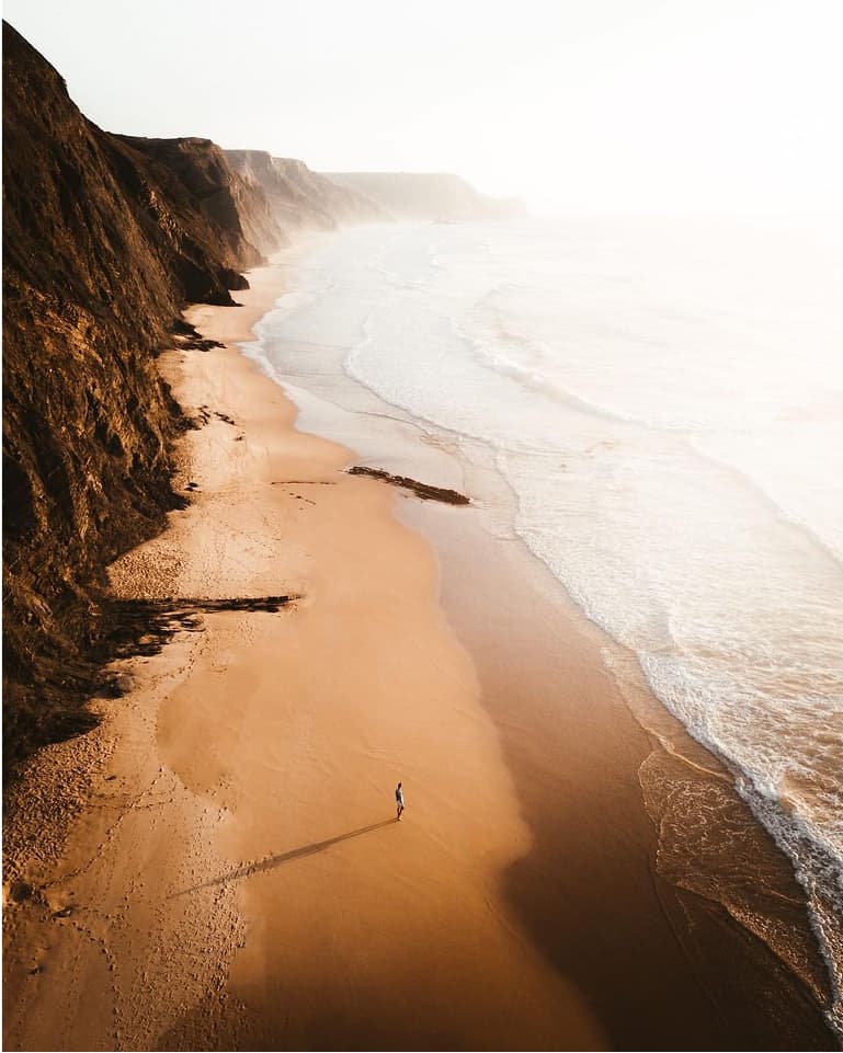 A person walks along a vast, empty beach with towering cliffs on one side and gentle waves on the other, under a bright, hazy sky.